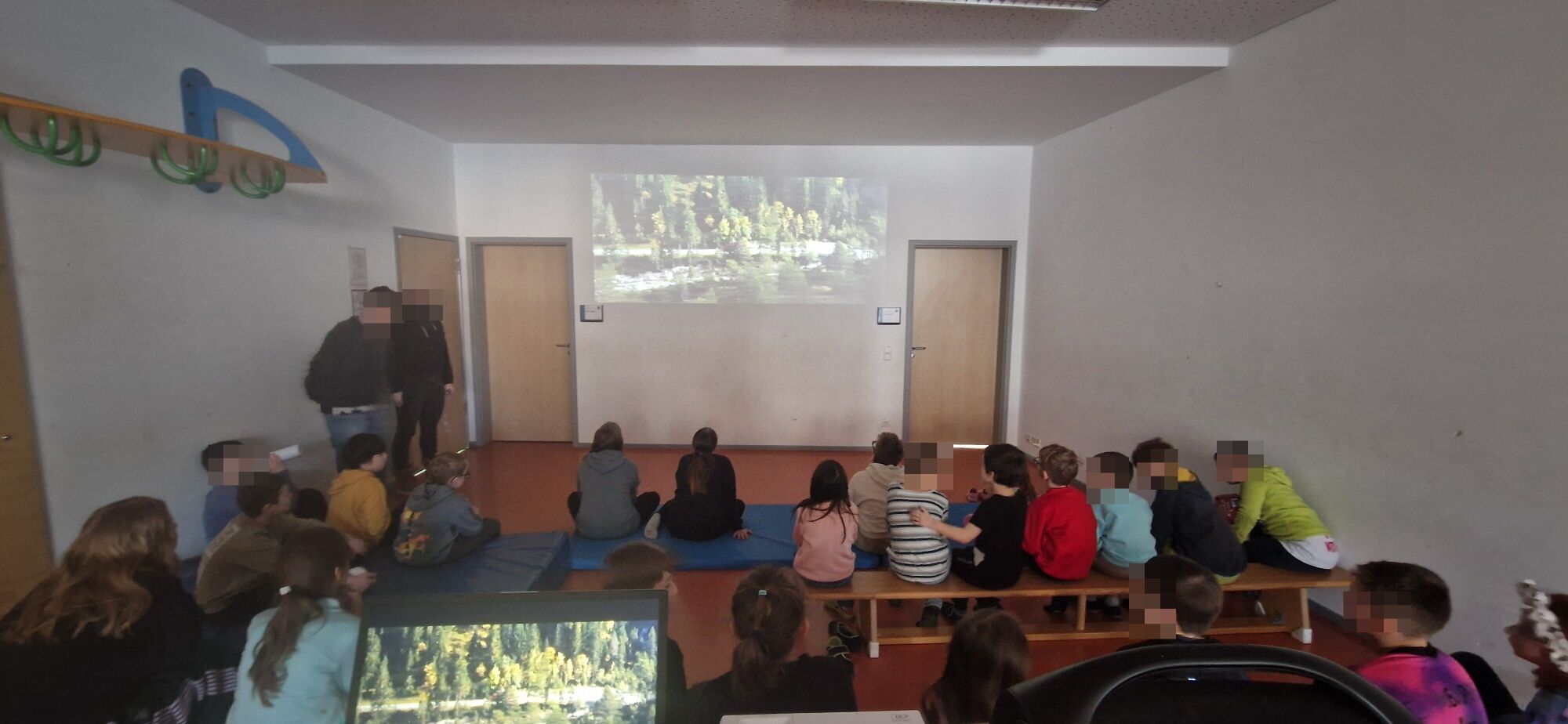 Viele Kinder sitzen auf B&auml;nken und schauen auf einen Monitor