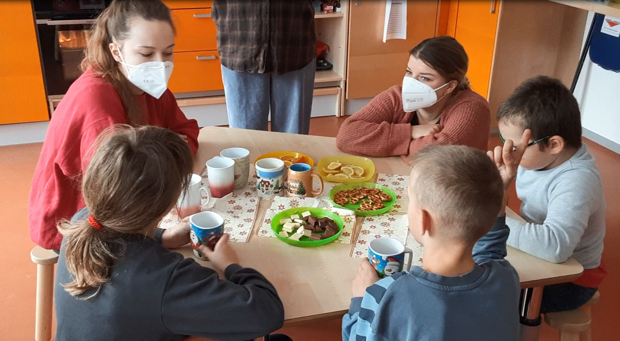 © SEB Leipzig Zwei Auszubildende sitzen mit drei Hortkindern an einem Tisch. Auf dem Tisch stehen Tassen und Teller mit Orangen, Zitronen, Schokolade und Brezeln