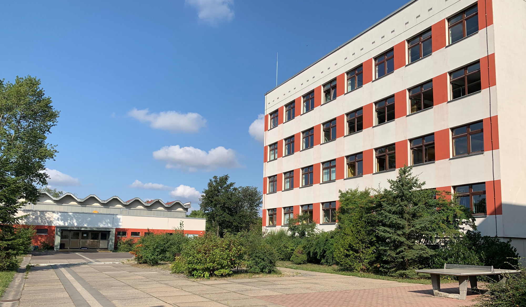 Vor einem großen weiß-reoten gebäude sind Pflanzen und eine Tischtennisplatte zu sehen.