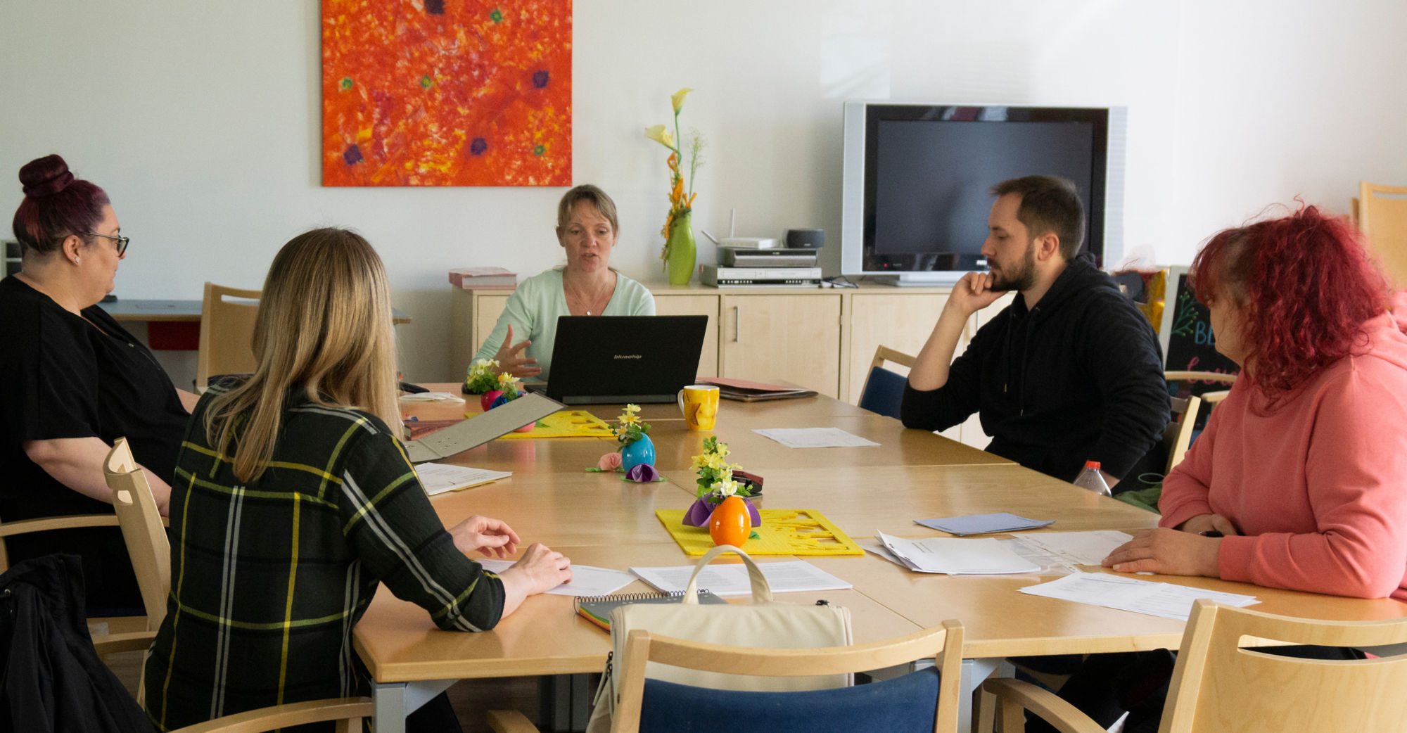 Fünf Menschen sitzen an einem großen Tisch mit Unterlagen und Laptop.