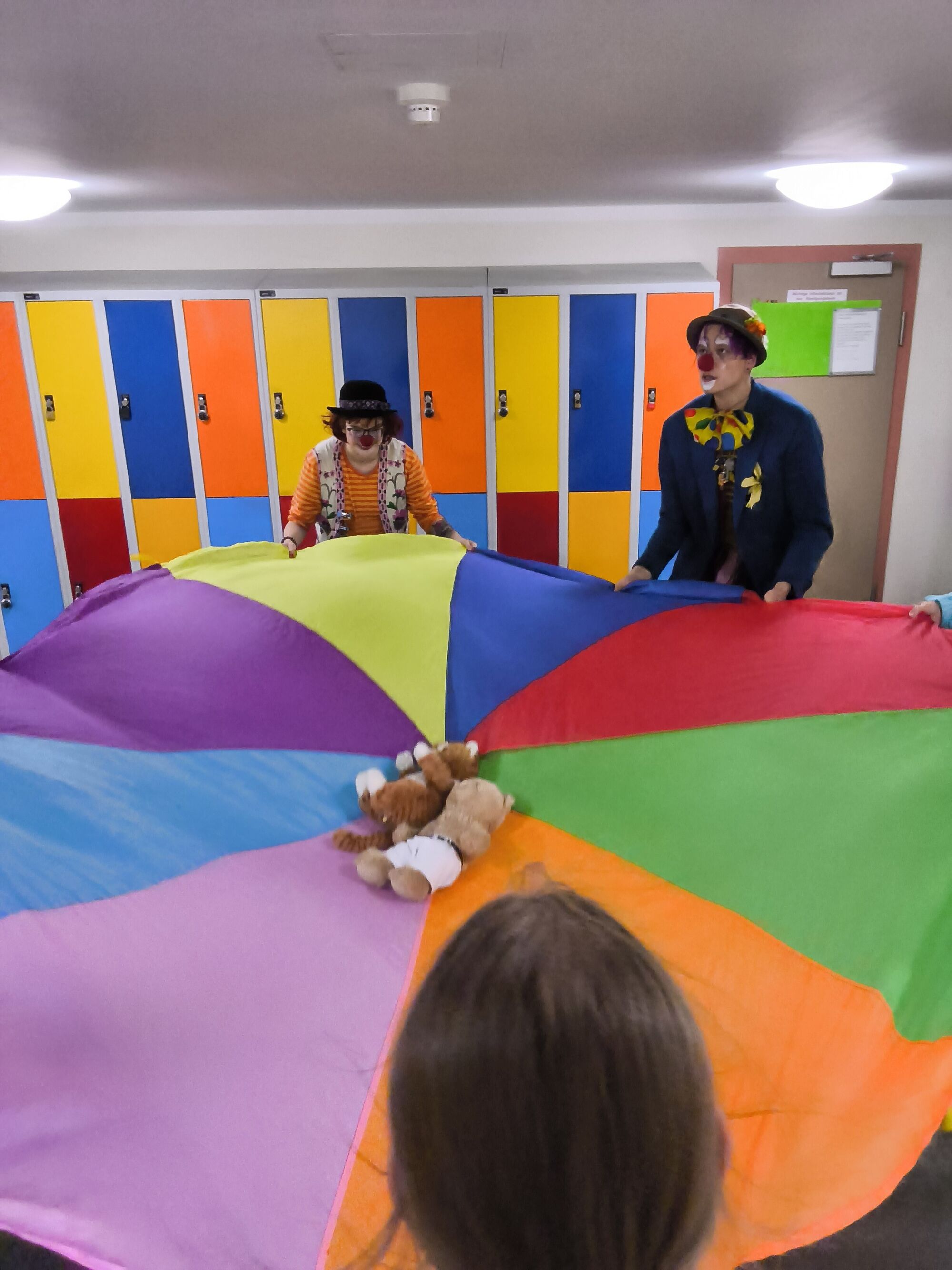 Zwei Clowns und Kinder spannen ein Schwungtuch. In der Mitte liegen Kuscheltiere
