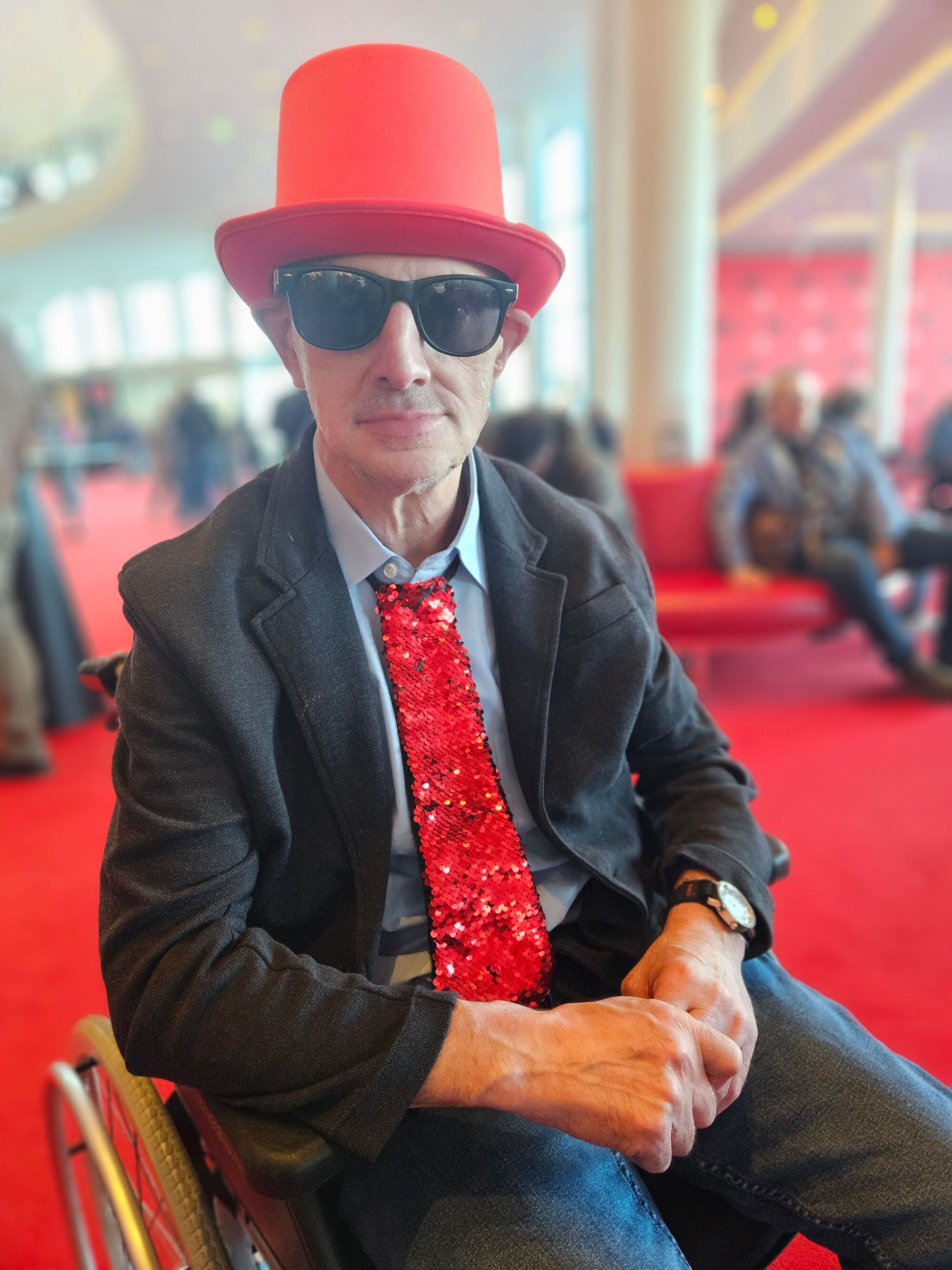 Ein Mann mit rotem Hut, roter Krawatte und Sonnenbrille im Foyer des Musicals