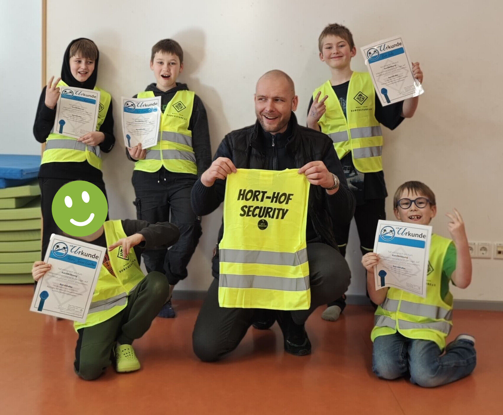 Kinder und ein mann hocken mit Urkunden und gelben Westen mit der Aufschrift "Hort-Hof-Security" auf dem Boden und lachen in die Kamera