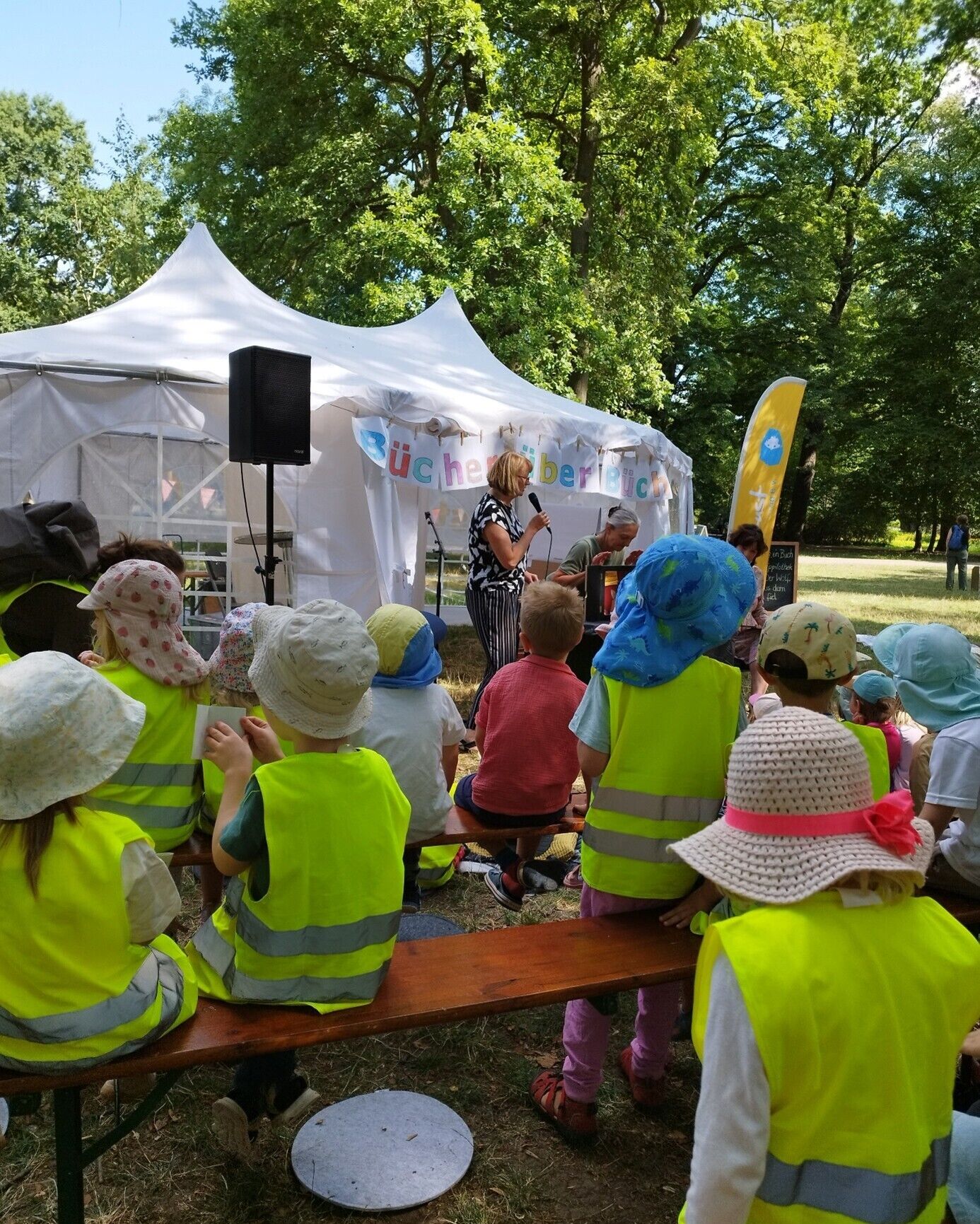 Kinder sitzen auf B&auml;nken in einem Park und h&ouml;ren einer Frau mit Mikrofon zu