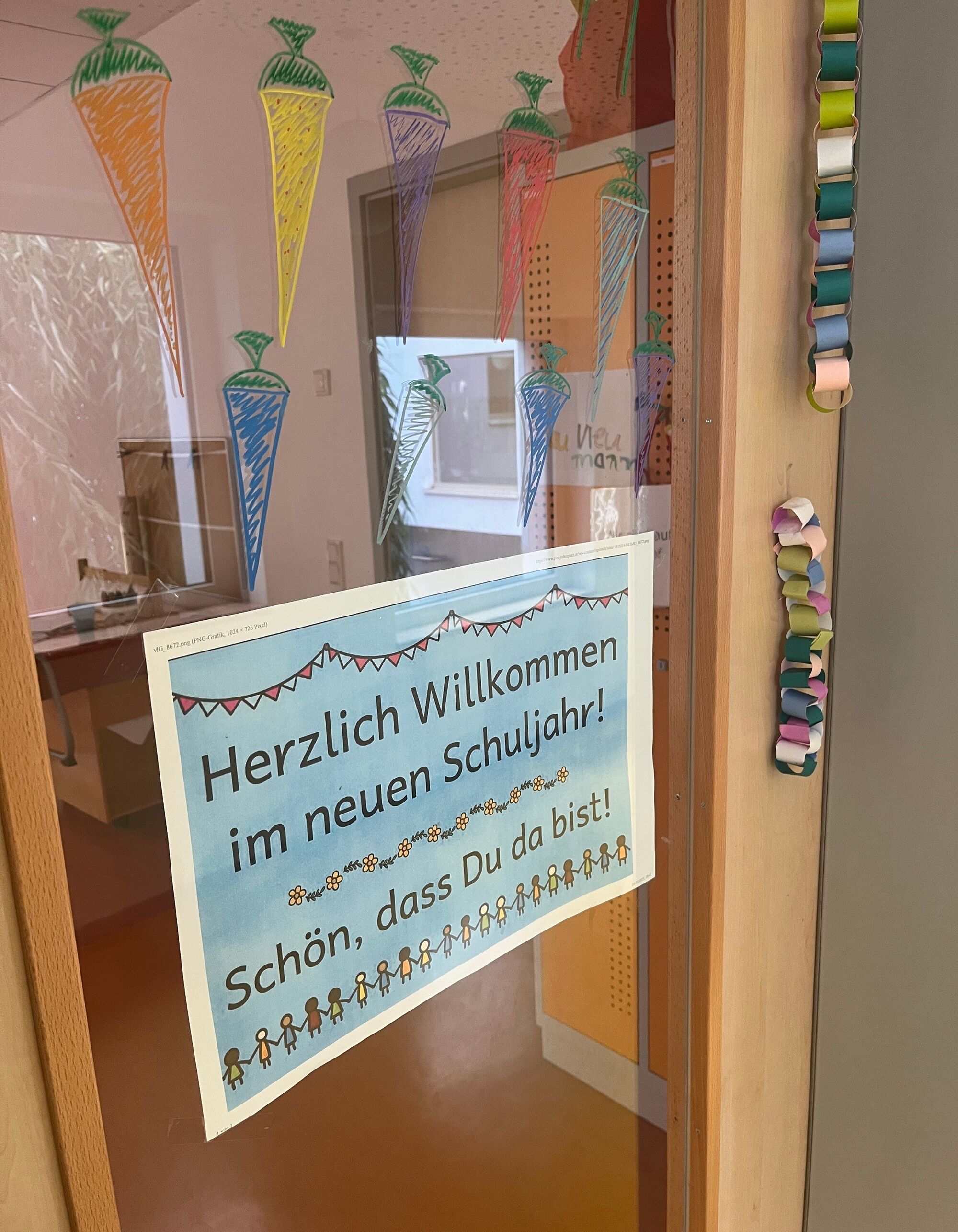 Ein Schild mit der Aufschrift "Herzlich Willkommen im neuen Schuljahr. Schön, dass du da bist." hängt an einer Tür. Die Scheiben sind mit bunten Schultüten bemalt.