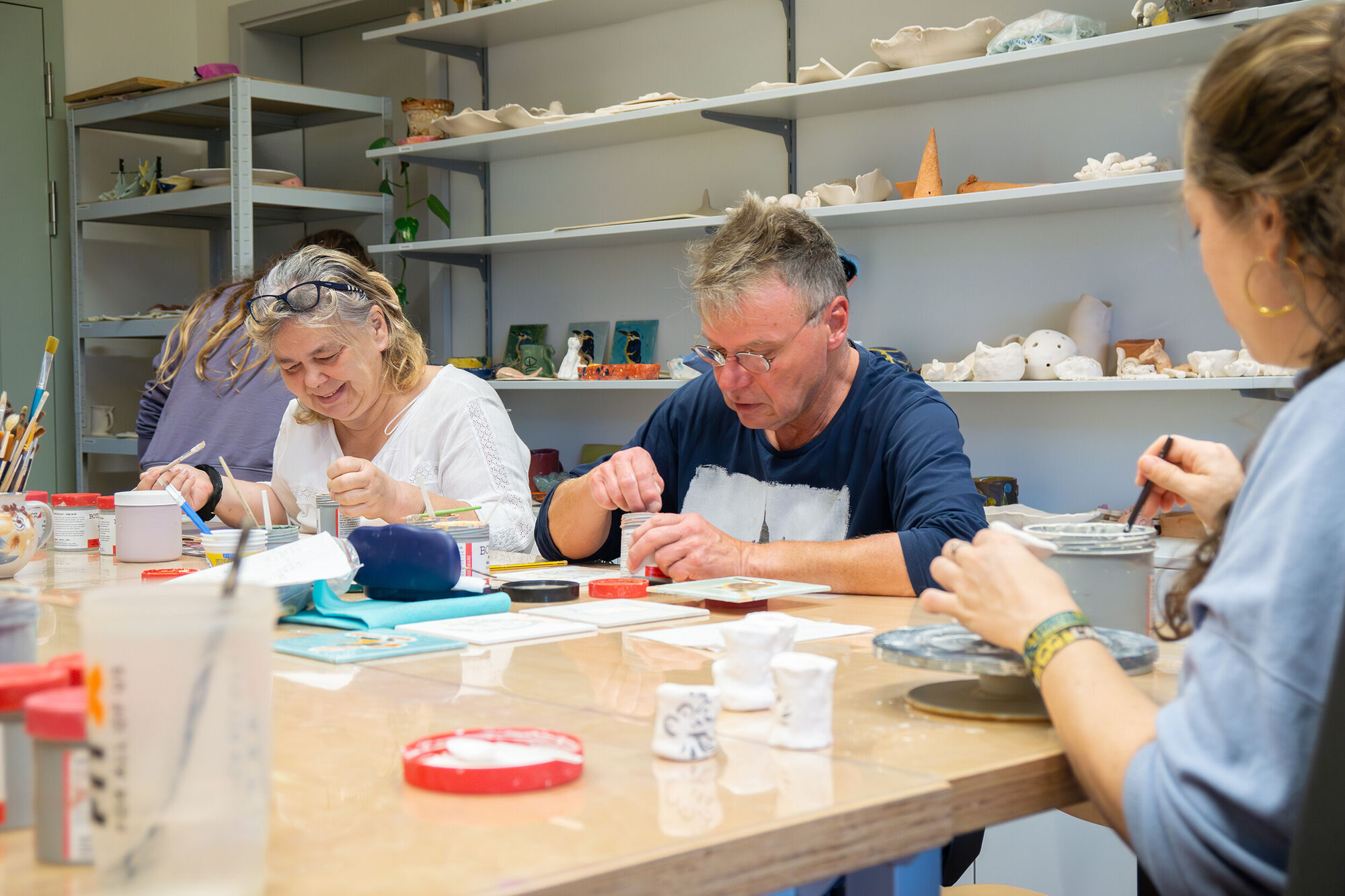 Menschen sitzen an einem großen Tisch mit Werkzeugen und arbeiten mit Ton. Im Hintergrund sind Regale mit fertigen Tonarbeiten zu sehen.