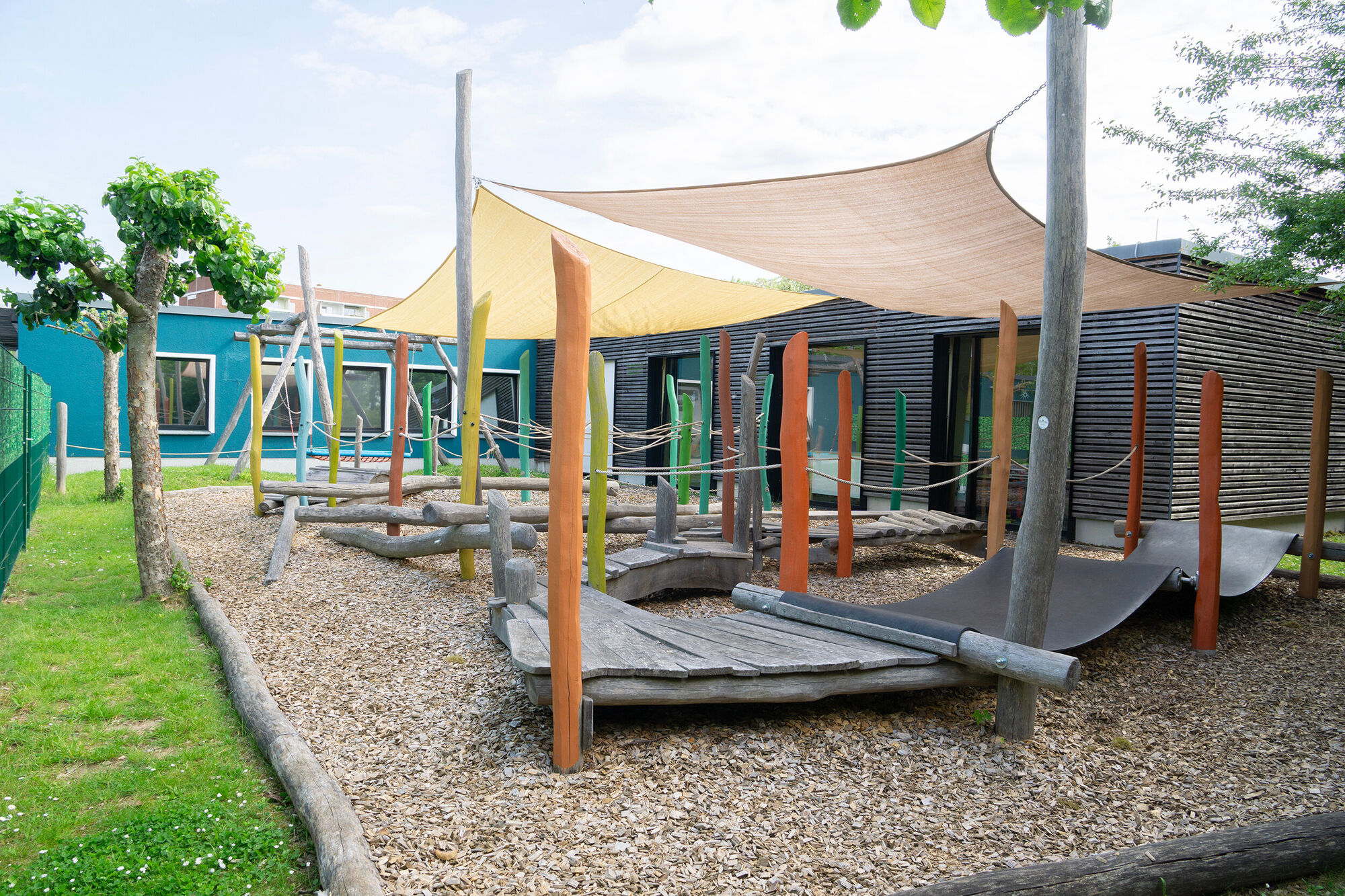 Möglichkeiten zum Klettern und Balancieren aus Holz und  Kunststoffmatten. Ringsherum ist Fallschutz angeschüttet. direkt daneben sind die Gebäude.