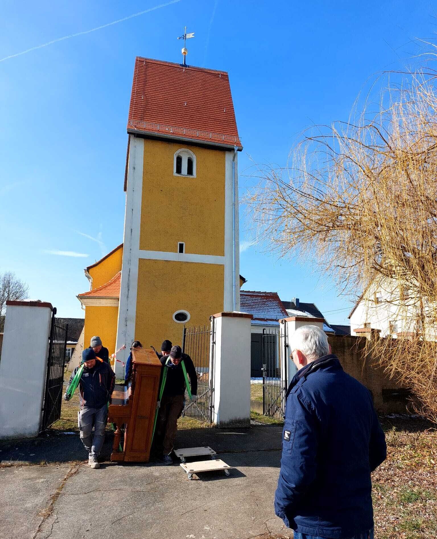 Ein Klavier wird von vielen Männern getragen. Im Hintergrund ist die Kirche zu sehen