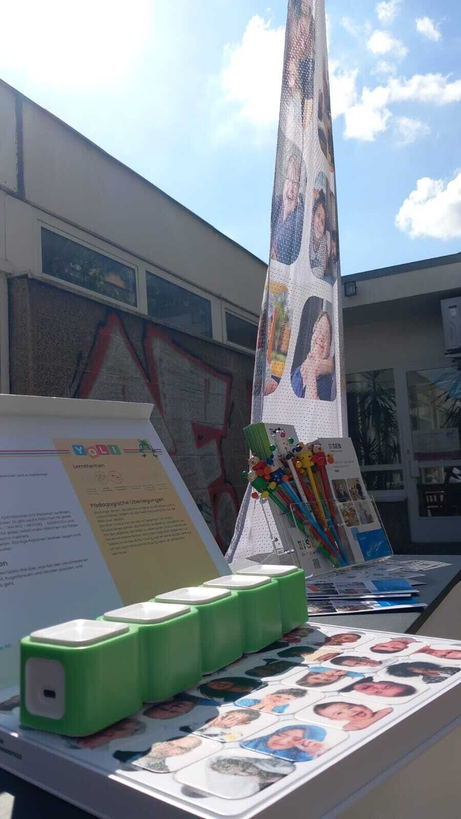 ein Tisch mit Yoli Spielset bestehend aus grünen Quadern und Bildern unterschiedlicher Menschen davor