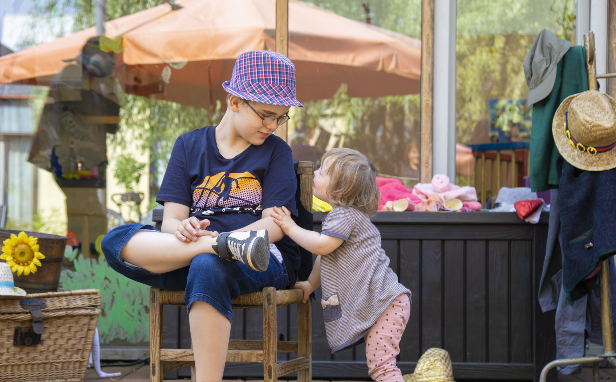 Ein kleines Mädchen zieht sich an einem Stuhl hoch, auf dem Ein Junge mit Hut sitzt und ihr zulächelt.