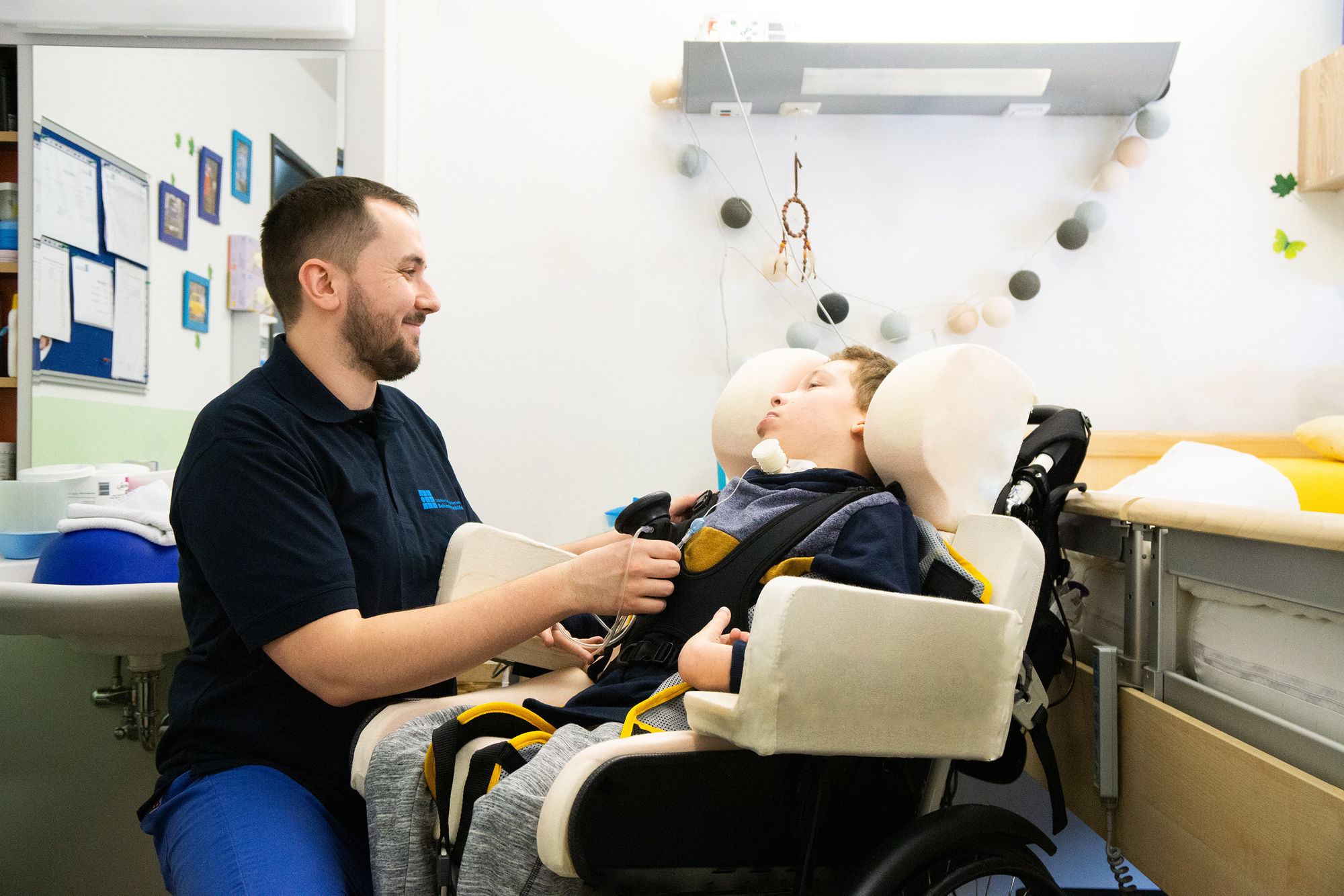 Ein Mitarbeiter prüft eine Trachialkanüle bei einem im Rollstuhl sitzenden Menschen