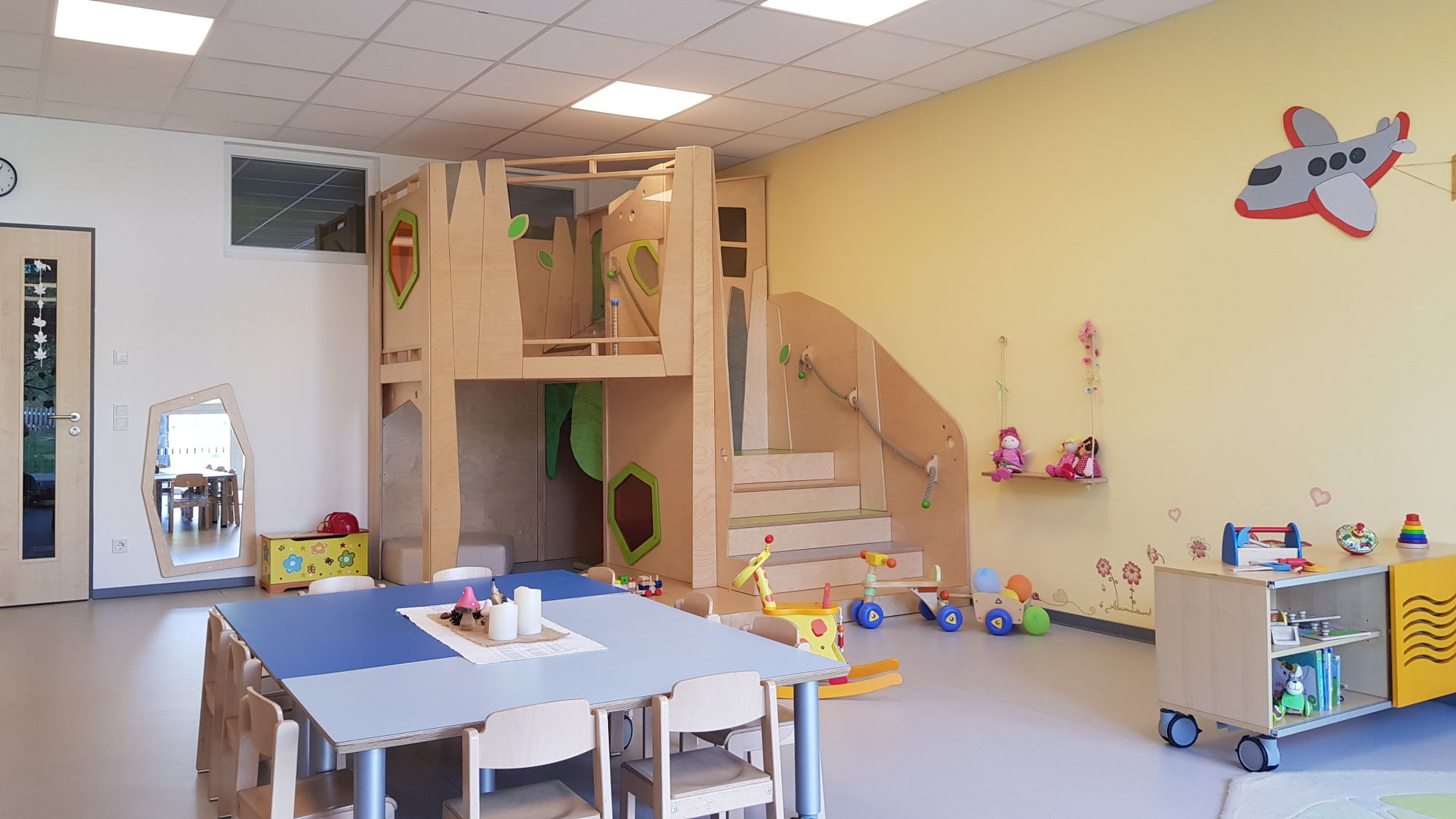 Ein Gruppenraum mit eingebauten Kletter- und Versteckmöglichkeiten, Rollkästen mit Spielsachen, Tischen und Stühlen.