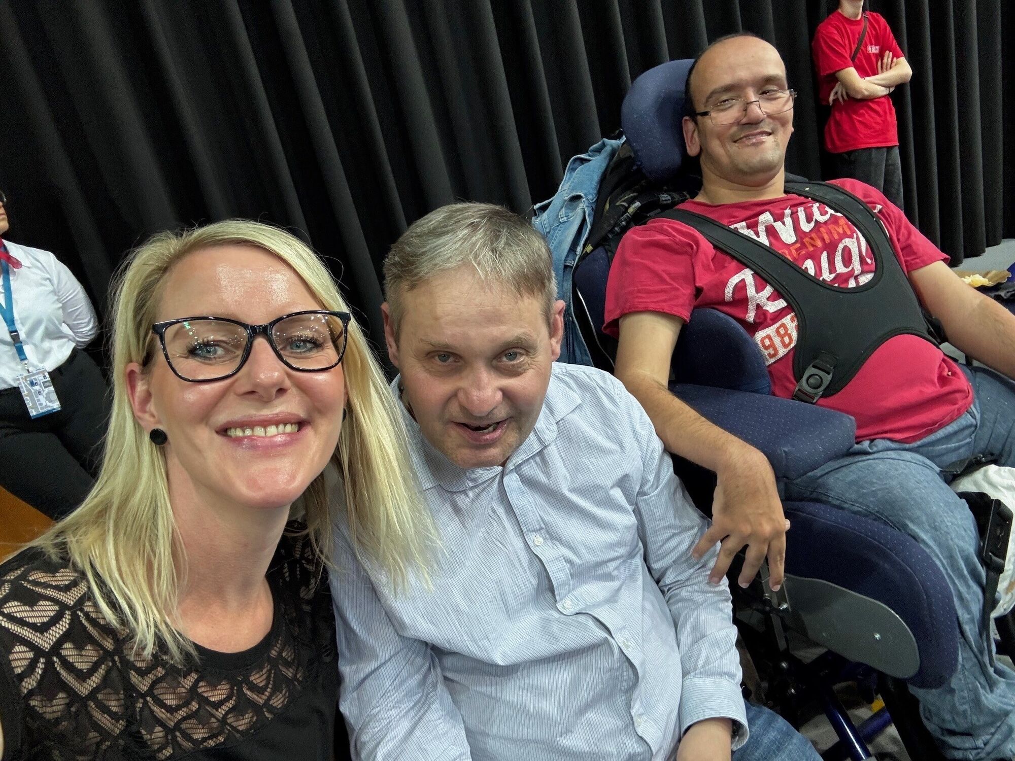 Ein Mitarbeiterin umarmt einen Bewohner. Daneben sitzt ein weiterer Bewohner im Rollstuhl. Alle lachen.