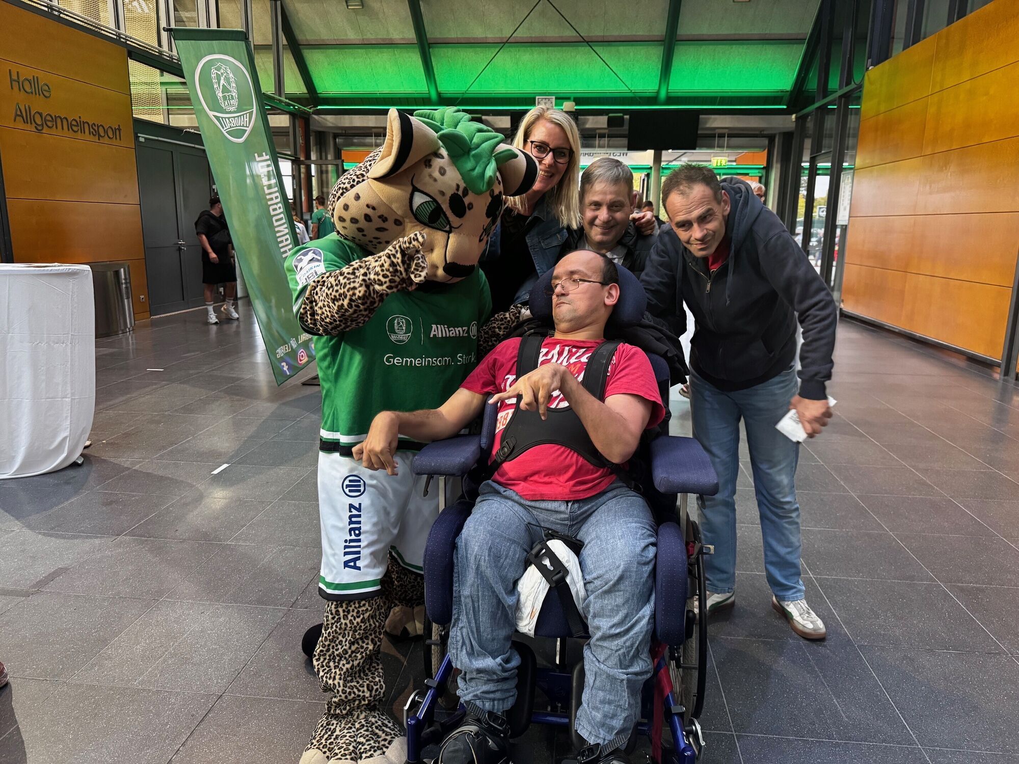 Vier Menschen stehen gemeinsam mit dem Maskottchen und lächeln in die Kamera. Einer von Ihnen sitzt im Rollstuhl.