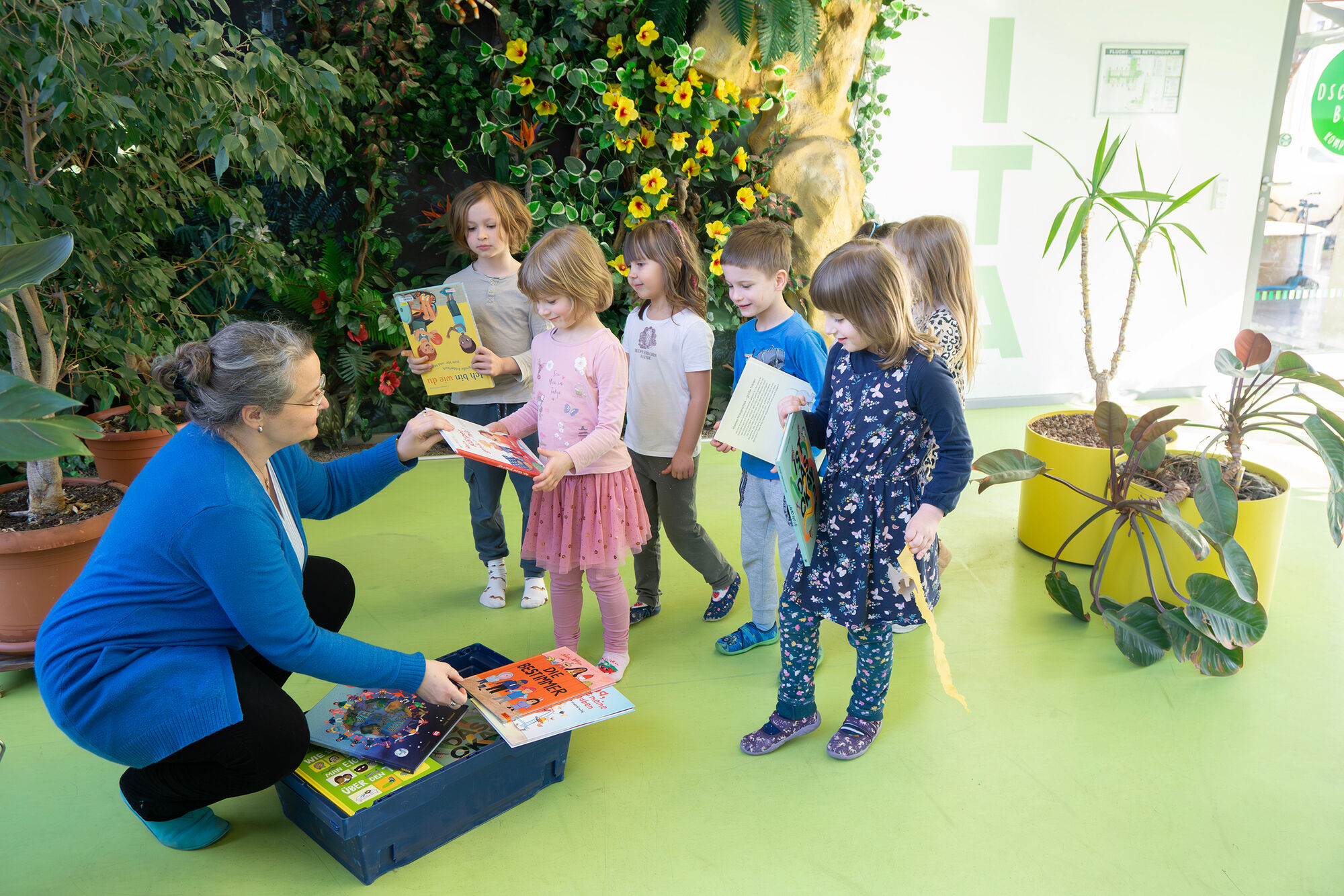 Ein l&auml;chelnde Frau hockt vor einer Kiste voller B&uuml;cher und &uuml;bergibt diese an lachende Kita-Kinder