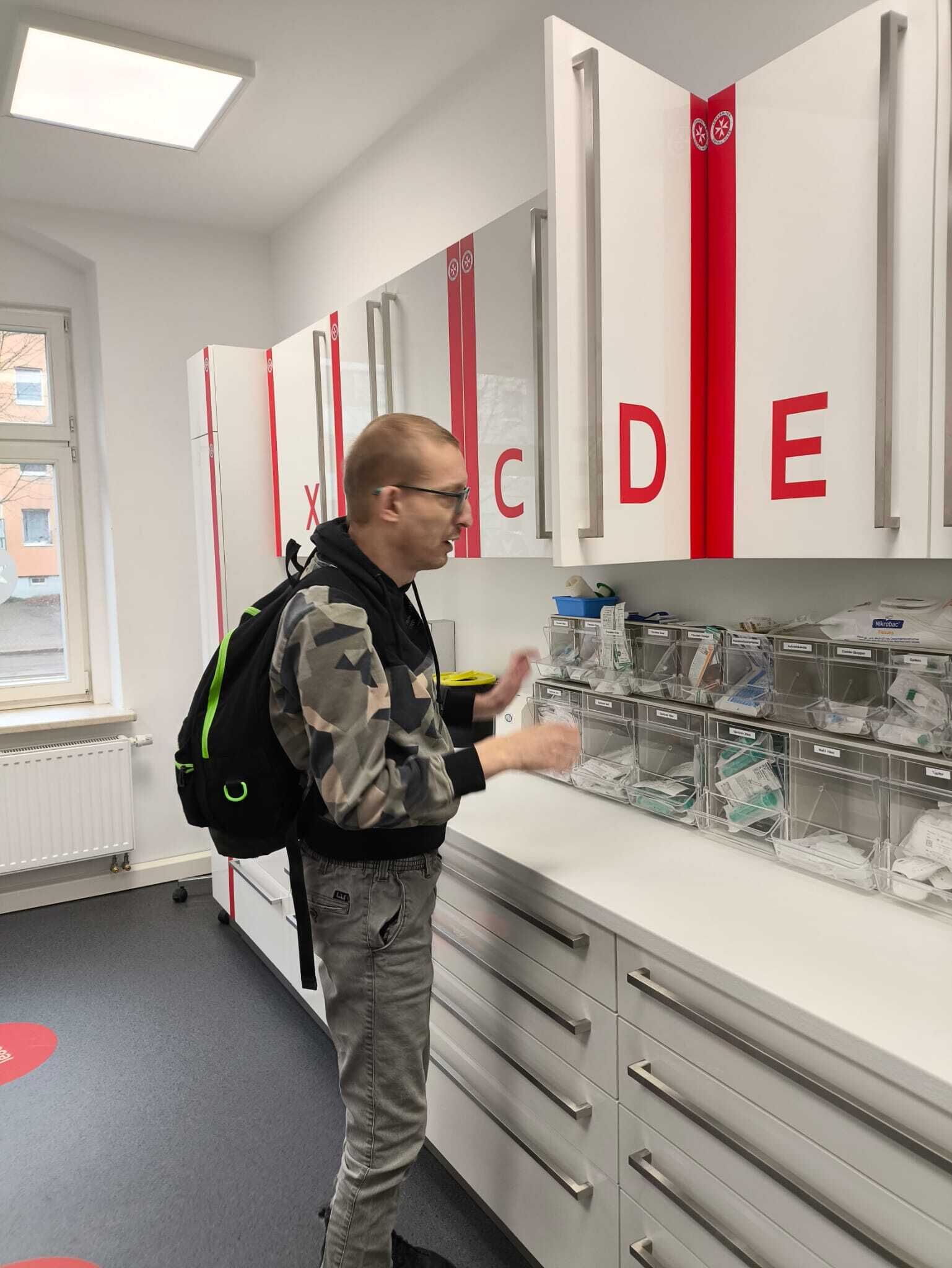 Ein Mann steht vor einem mit Buchstaben beschrifteten Schrank und vielen F&auml;chern mit Medikamenten