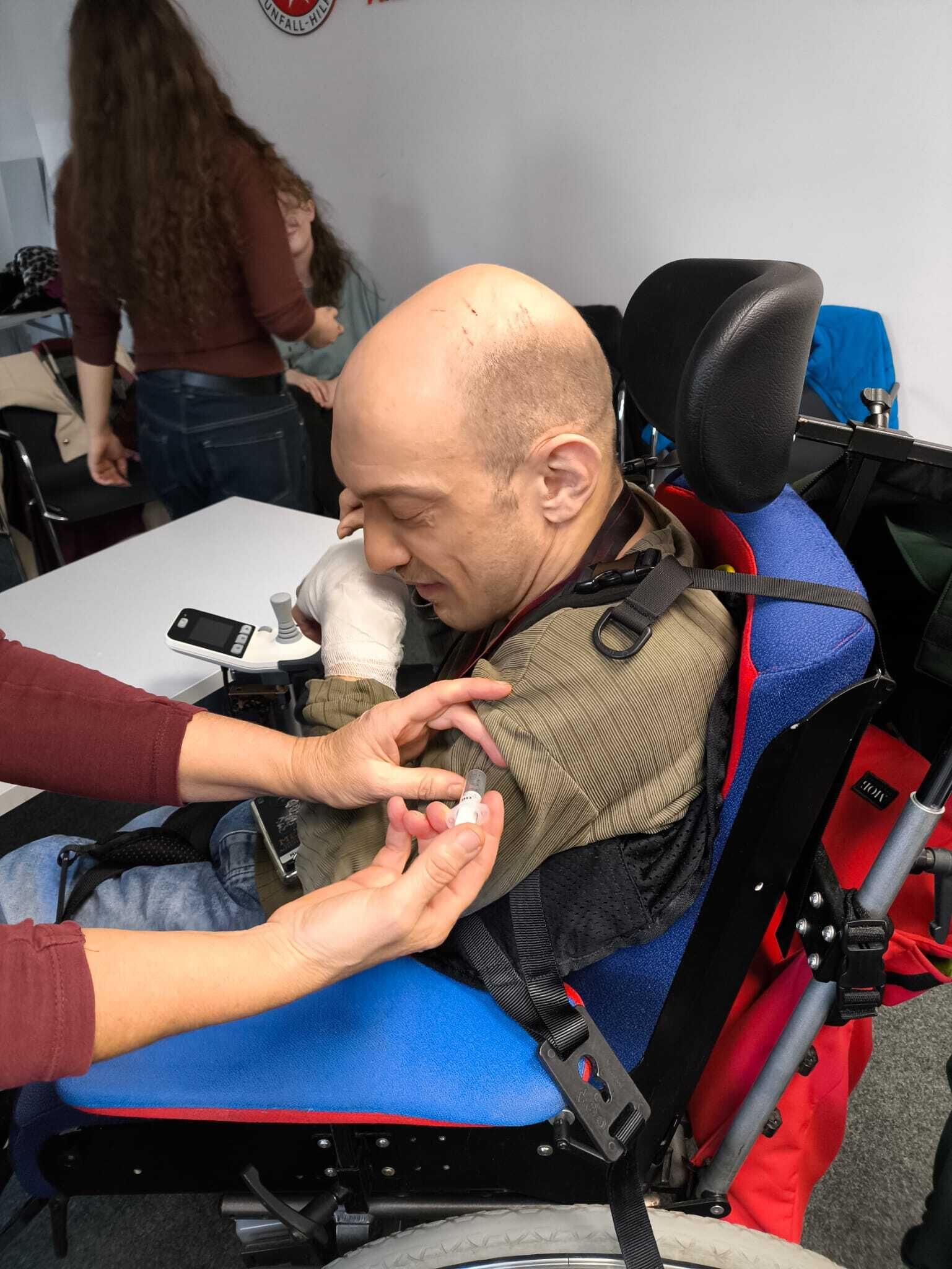 Ein Mann im Rollstuhl hat ein verbundenes Handgelenk. Es wird so getan als w&uuml;rde er eine Spritze erhalten.